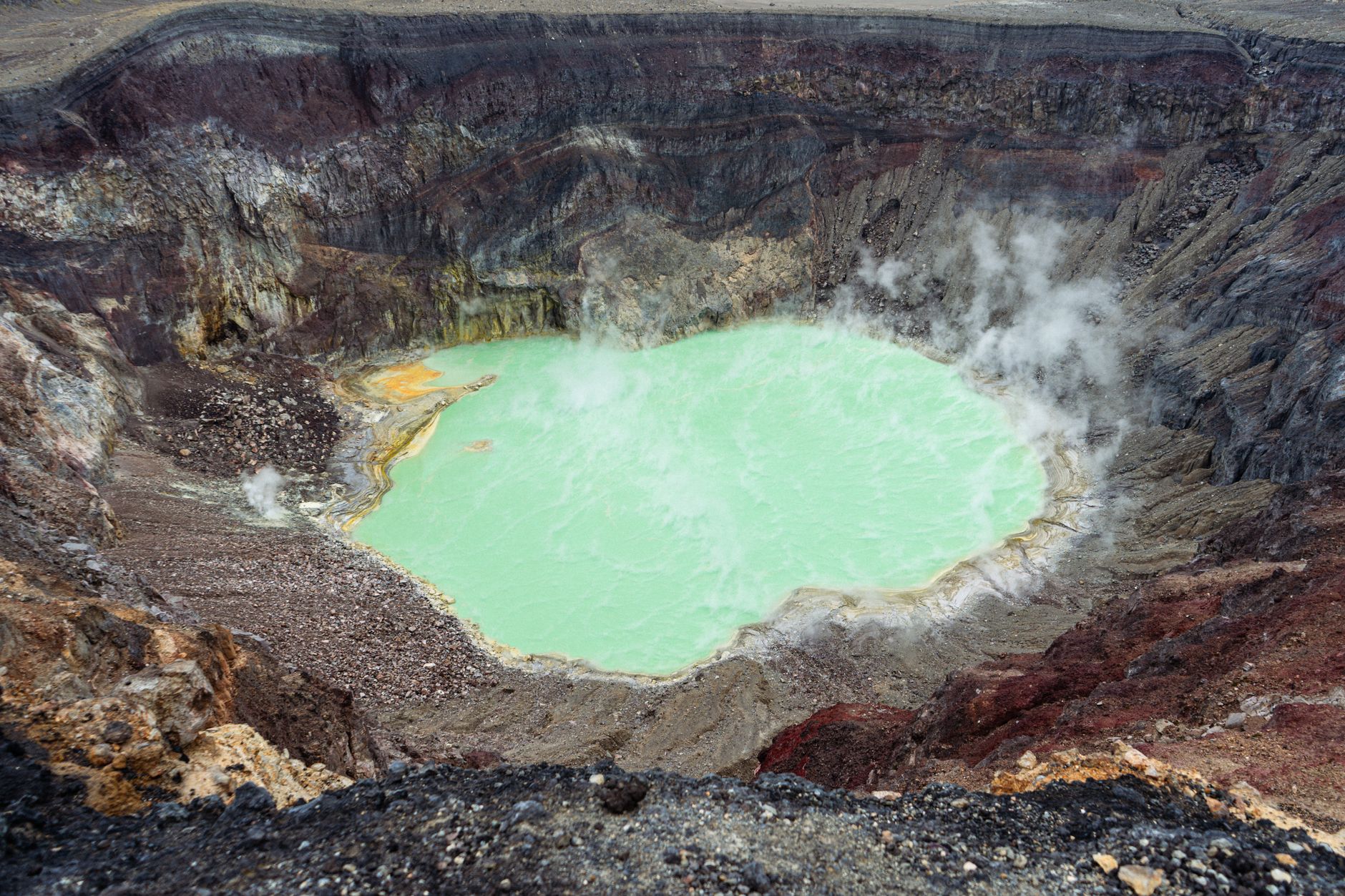 Aerial view of a steaming volcanic crater with a vivid turquoise lake and rugged rock formations.