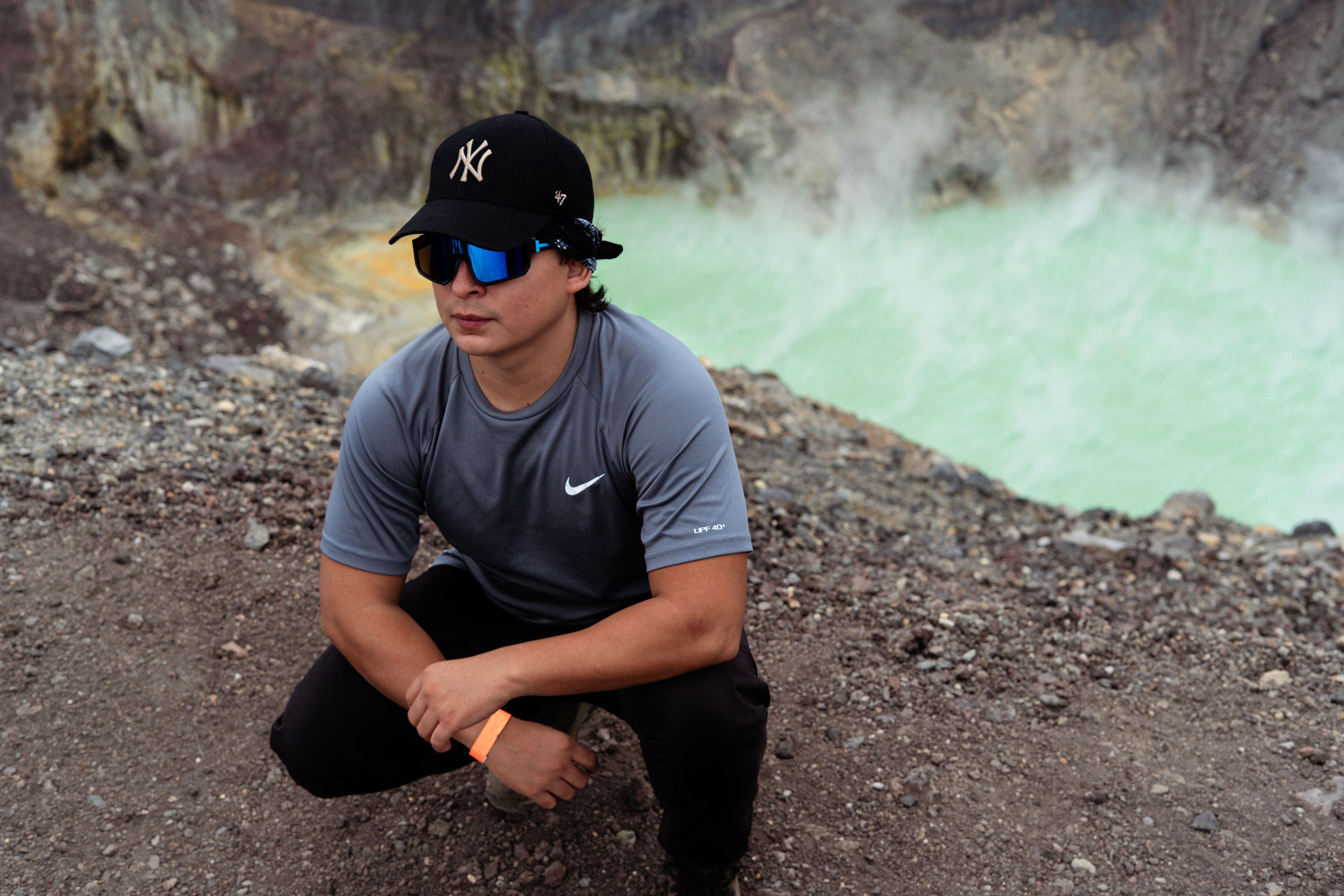 Young man wearing sunglasses squatting by a steaming volcanic crater, showcasing adventure and exploration.