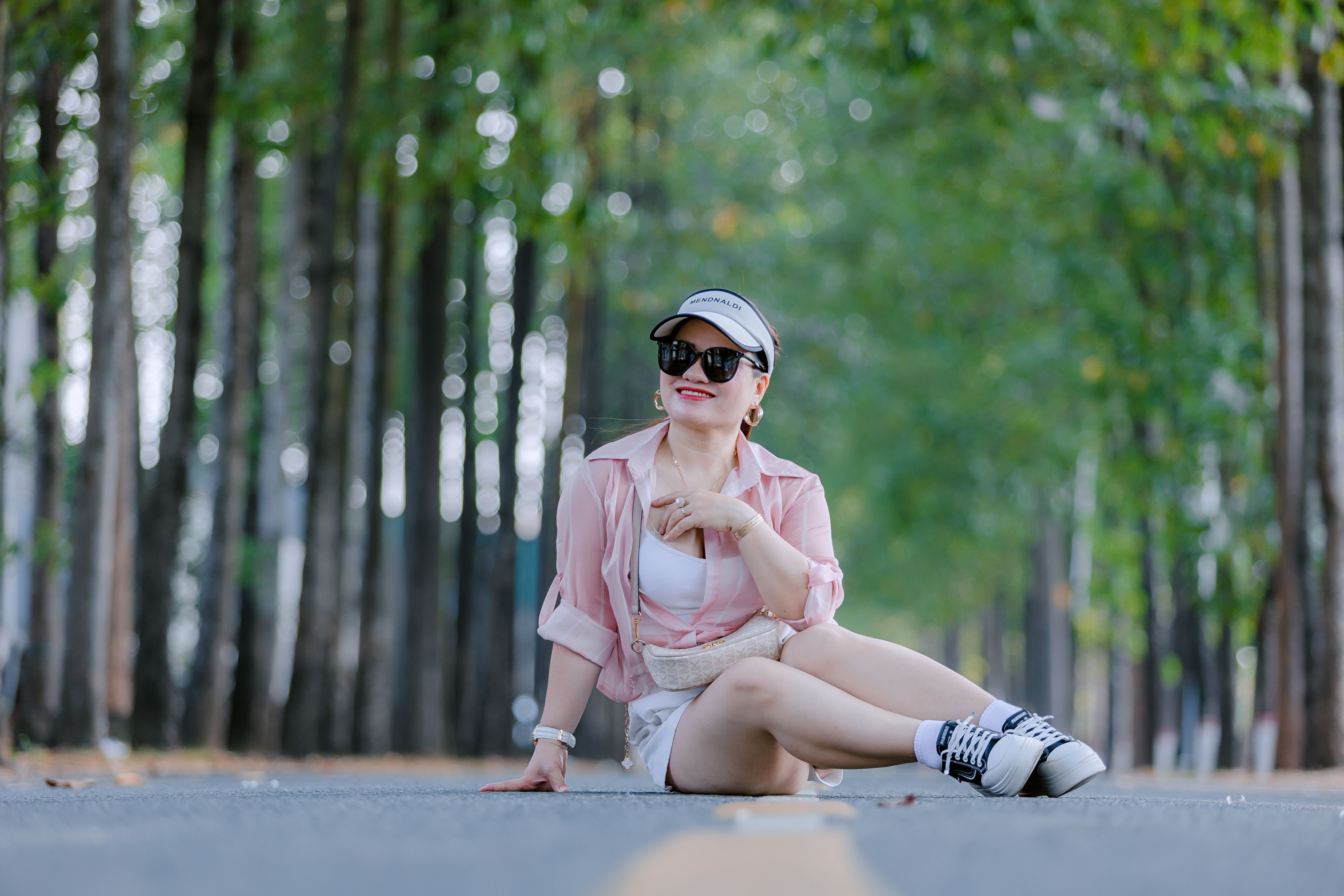 Fashionable woman in pink shirt sits on road lined with trees, enjoying a sunny day.