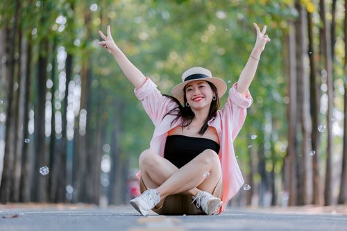 Free Smiling woman sitting cross-legged on a scenic tree-lined road, wearing a hat and raising peace signs. Stock Photo