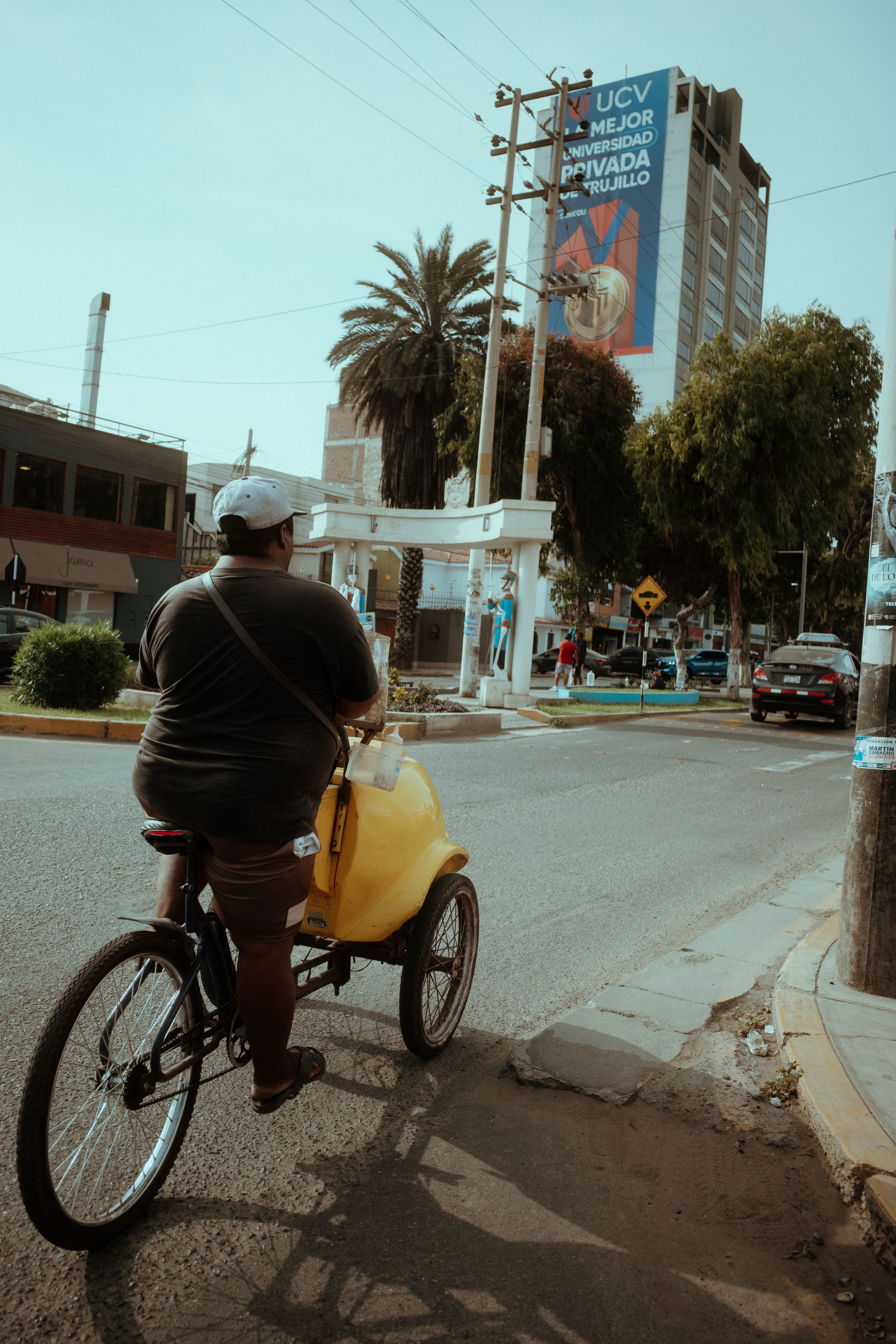 A rear view of a man cycling on an urban street in Trujillo, Peru, with buildings and greenery.
