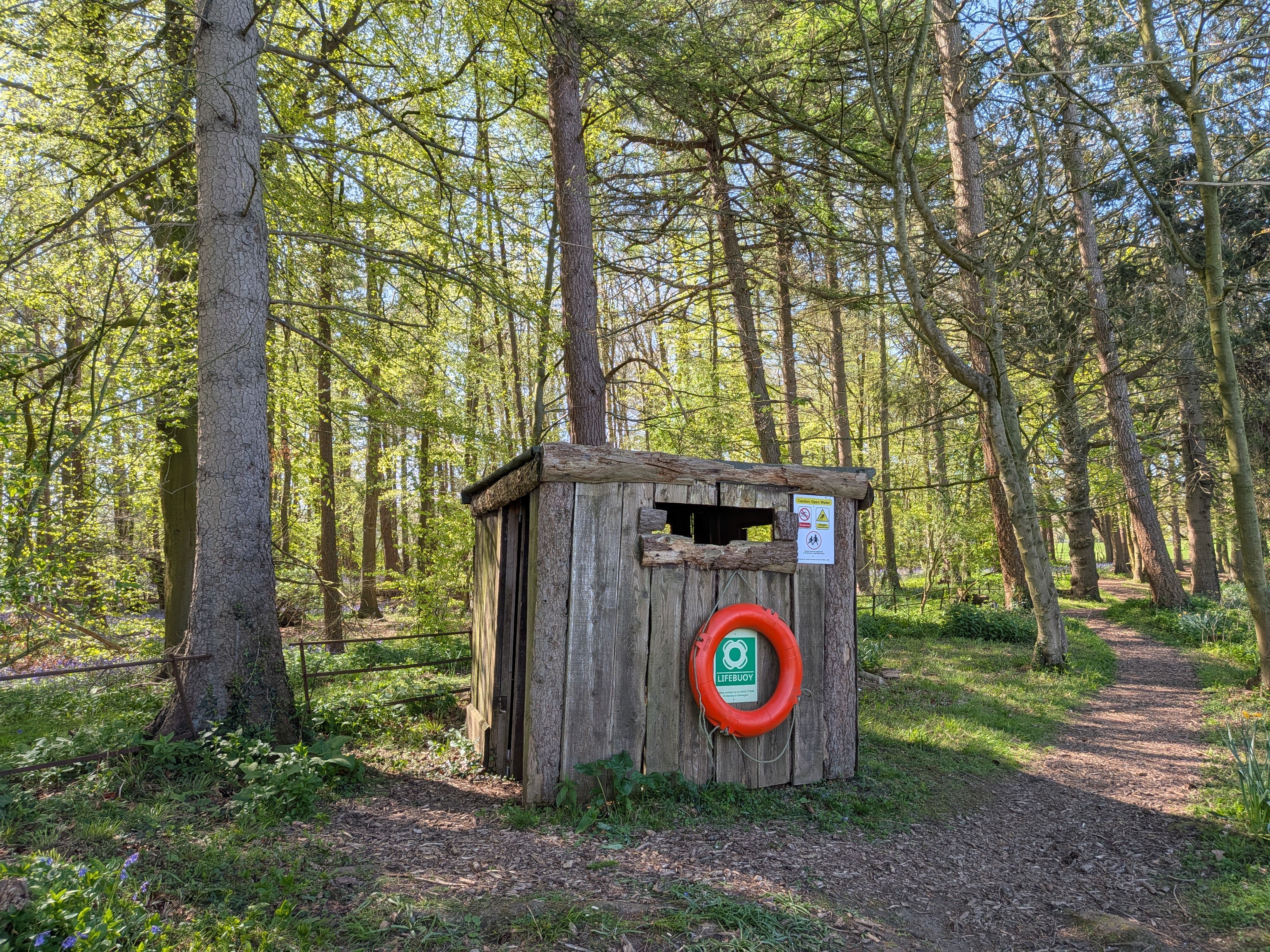 A rustic wooden cabin with a lifebuoy in a sunlit English forest, surrounded by trees.