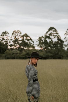 日落时分，一位身着条纹连衣裙、头戴帽子的优雅女子，静立在宁静的田野上