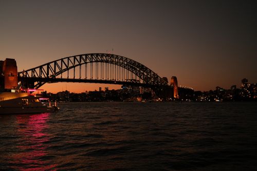 Free A stunning view of Sydney Harbour Bridge silhouetted by sunset, featuring a boat illuminated with vibrant lights on the water. Stock Photo