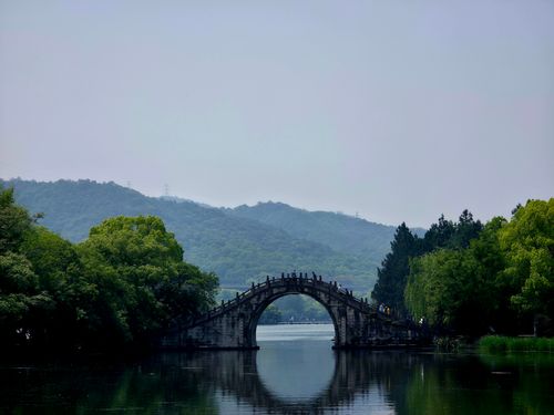 Free Serene landscape featuring a traditional stone arch bridge over a calm lake, with lush greenery and distant mountains. Stock Photo