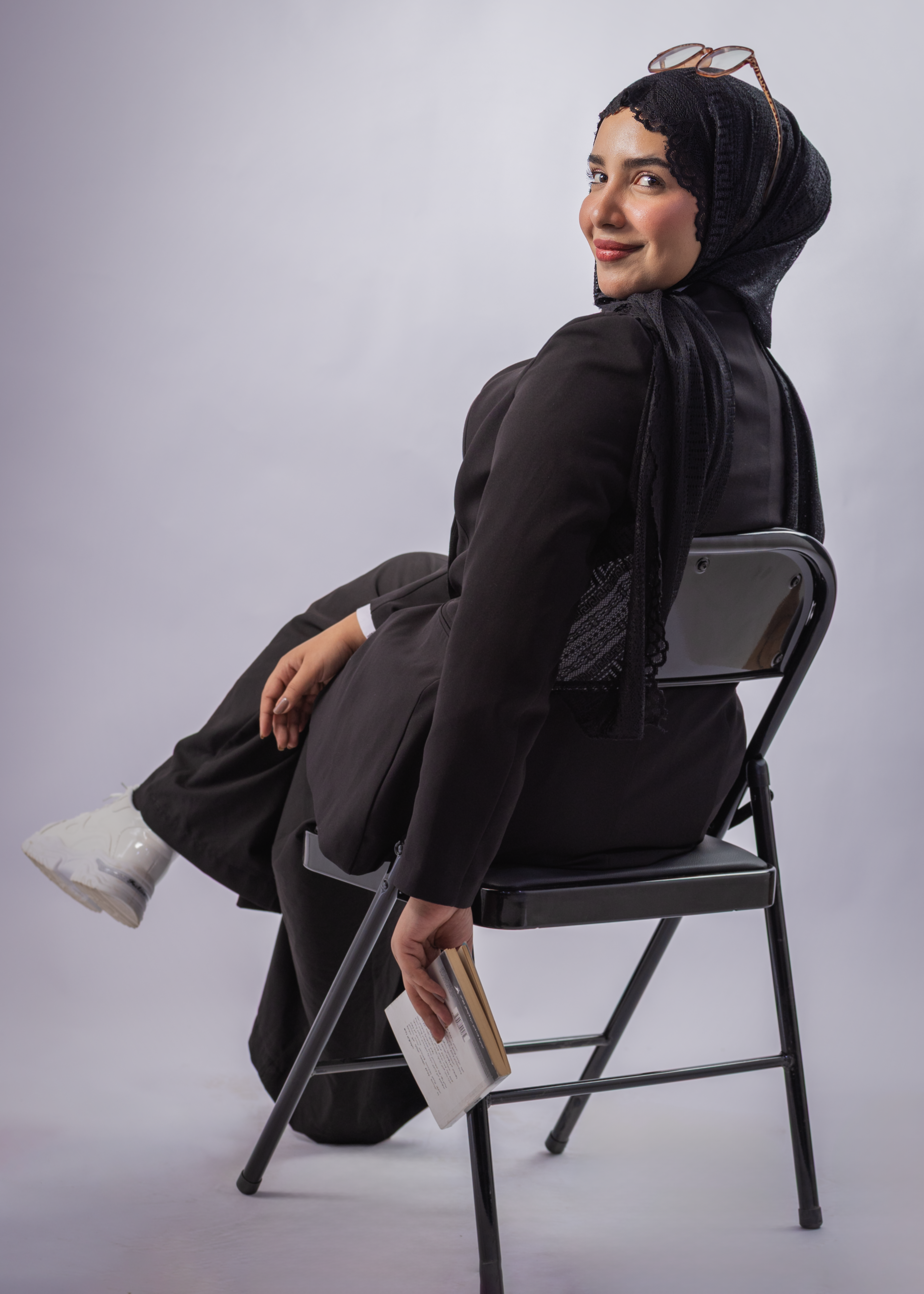 Smiling woman in hijab seated casually on a chair, holding a book.