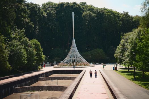 People walk near a tall modern sculpture in a lush green park on a sunny day.