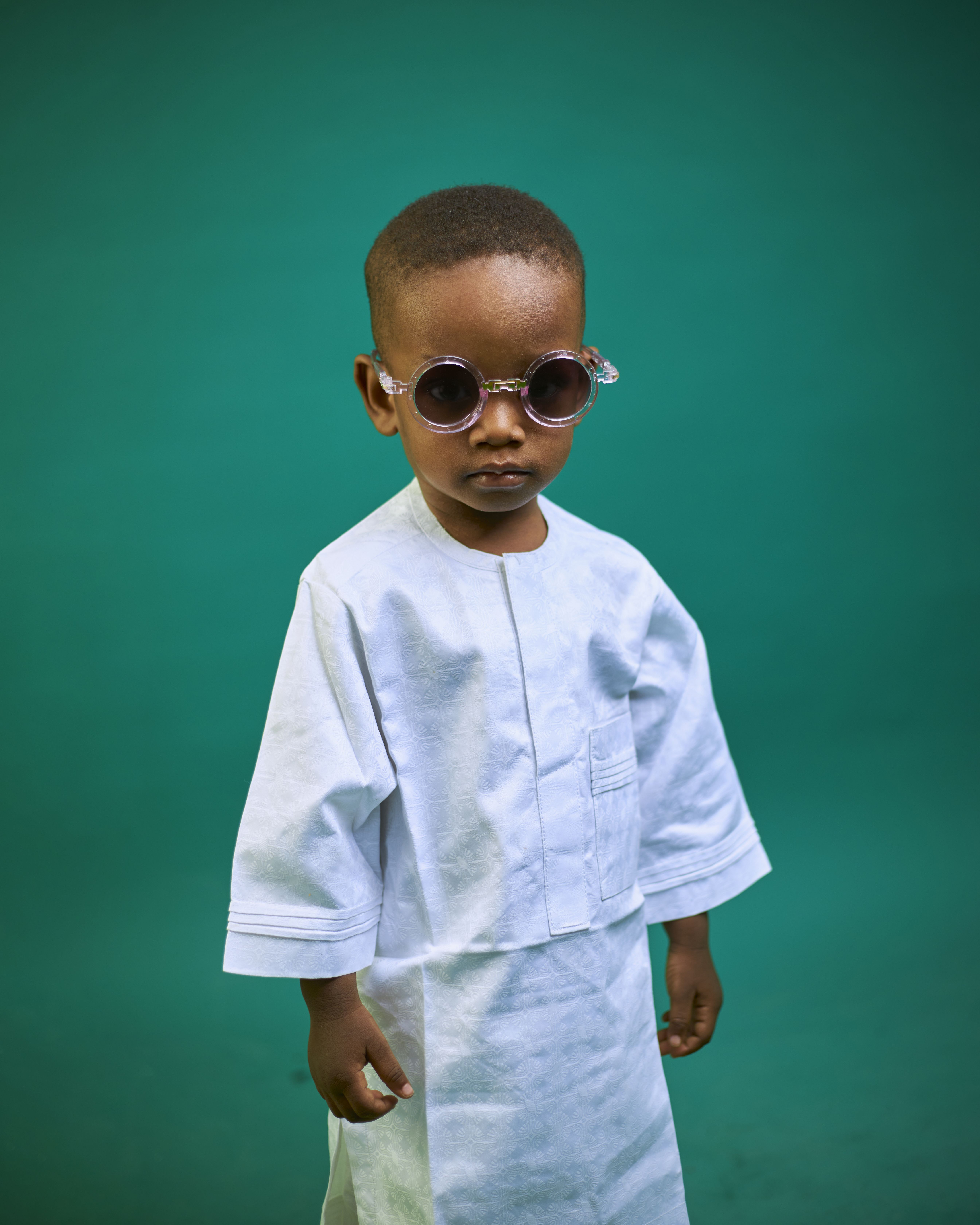 Portrait of a young boy wearing traditional attire and sunglasses against a green background.