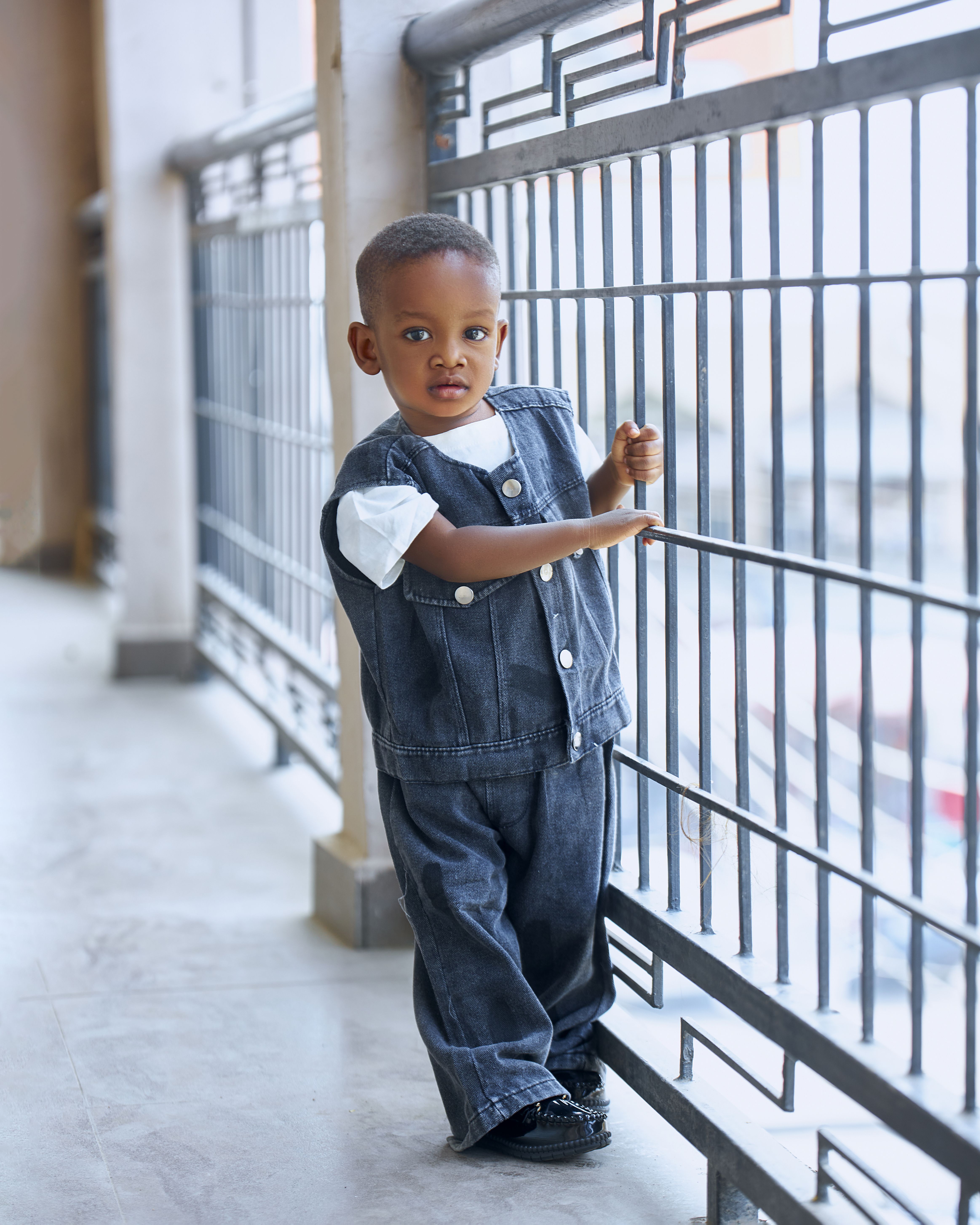 Cute toddler in denim vest and jeans standing on a balcony.
