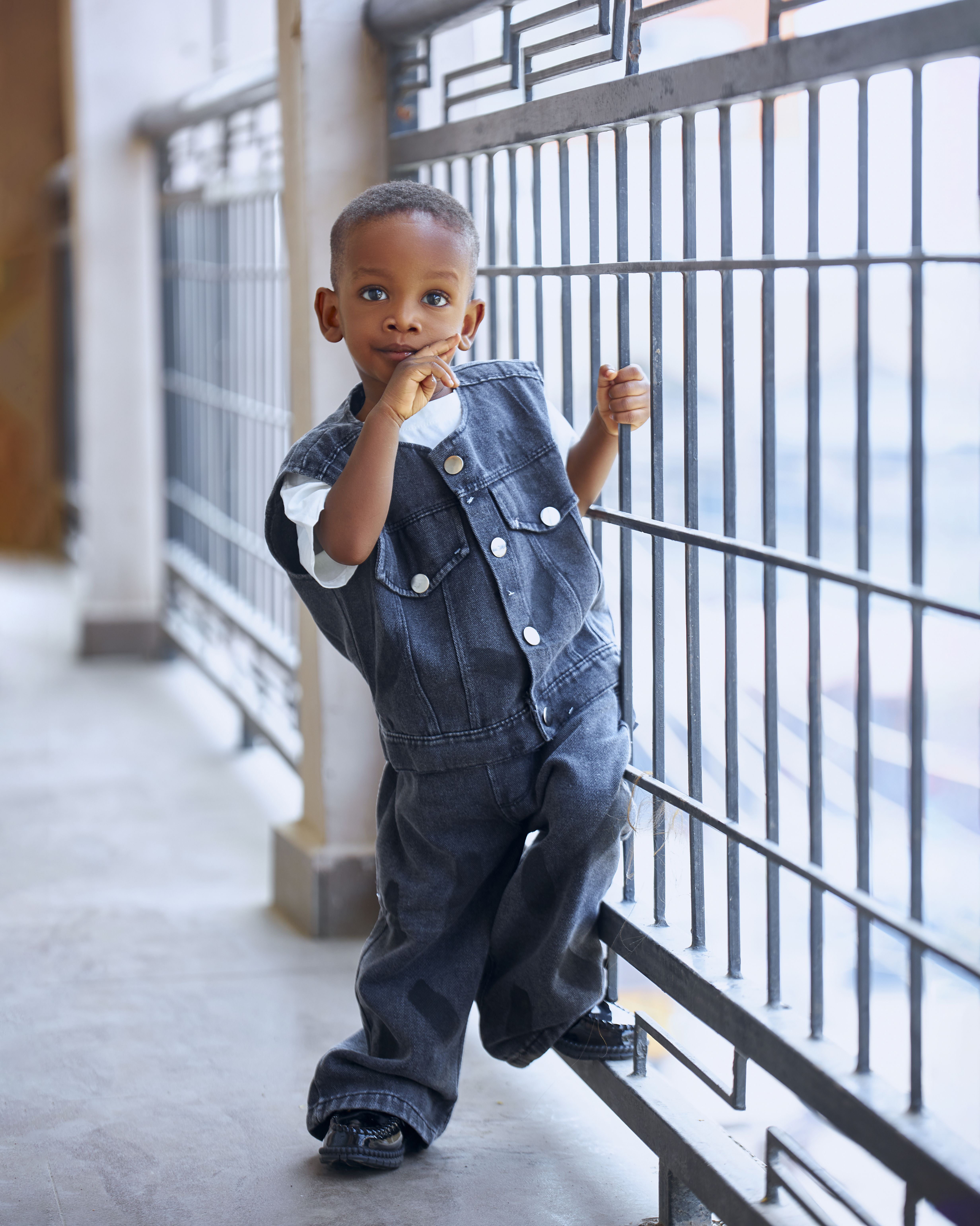 A young child in a denim outfit poses playfully against a railing, indoors.