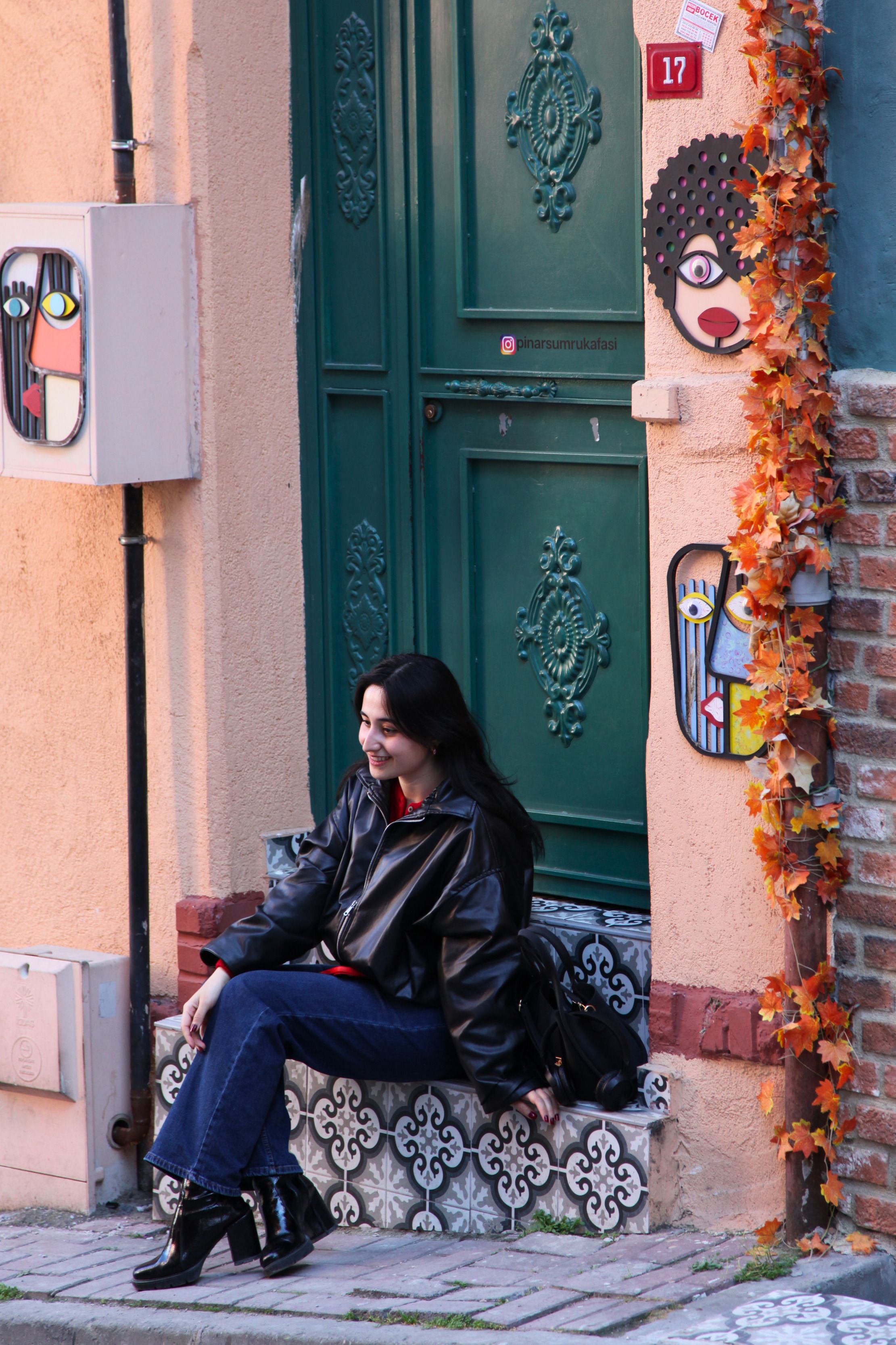 A woman in a stylish outfit sitting by a decorated wall with autumn leaves and artwork, embracing fall vibes.