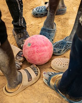 Children's feet encircle a worn pink soccer ball on sandy ground, showcasing play and community.