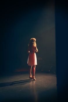 A woman standing on stage, illuminated by a spotlight, wearing a light dress.