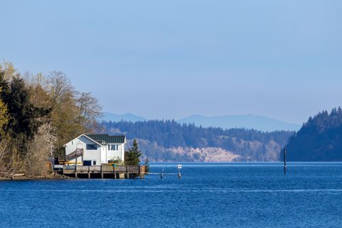 Free A serene lakeside house surrounded by trees with distant mountains under a blue sky. Stock Photo