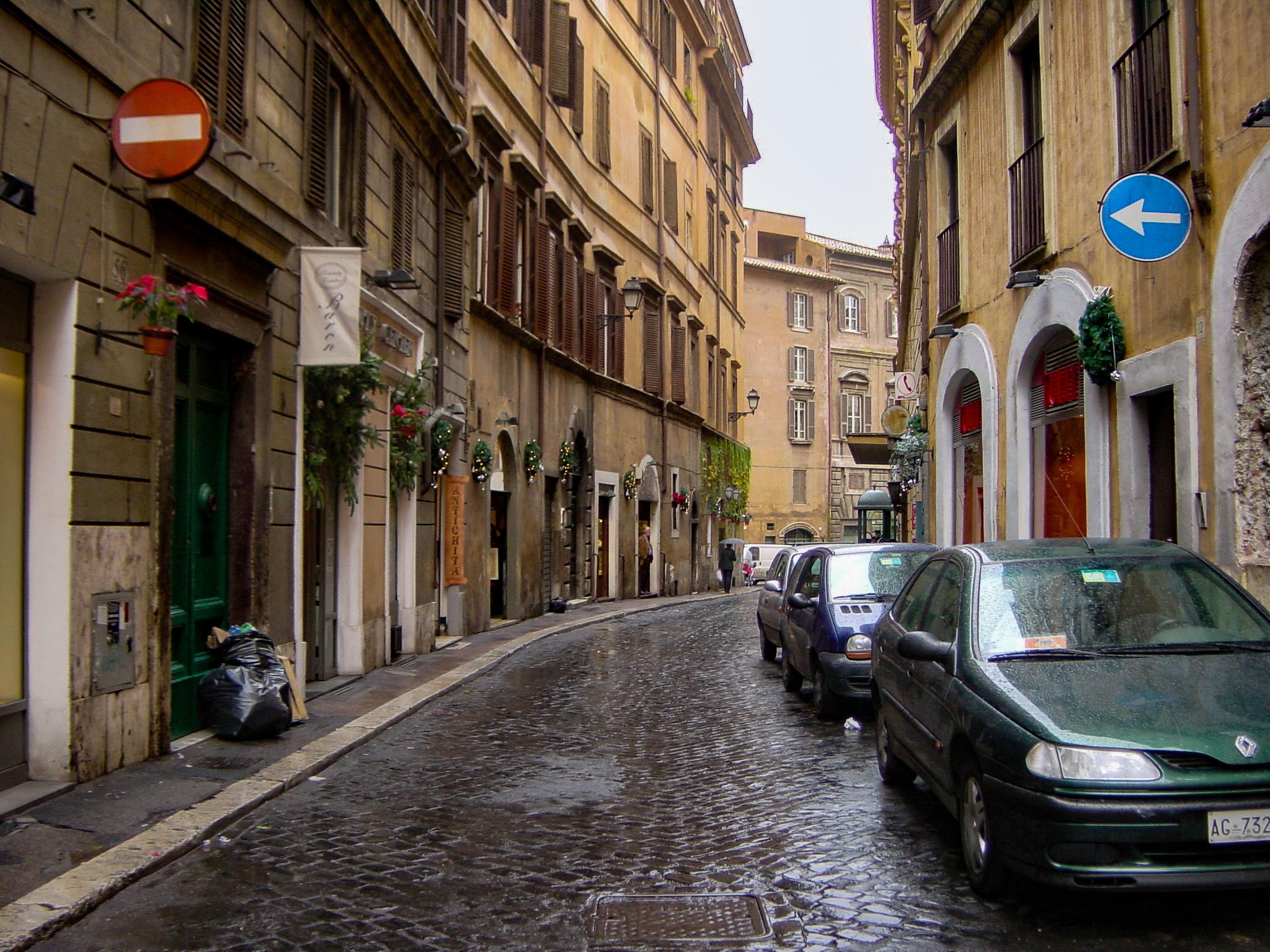 Charming, rainy cobblestone street in Rome, lined with historic buildings and parked cars.