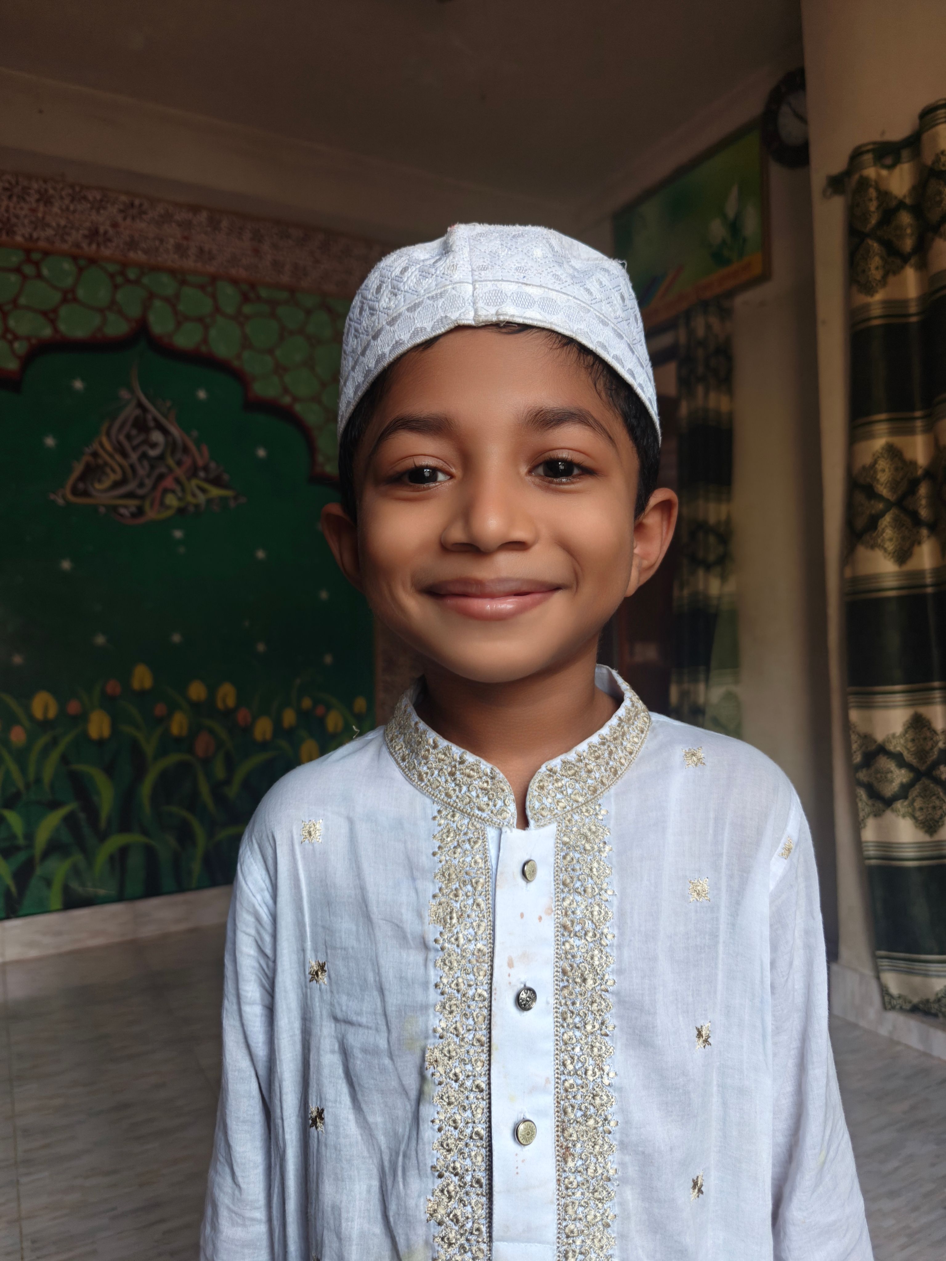 Young boy in traditional white attire smiling indoors with ornate background.