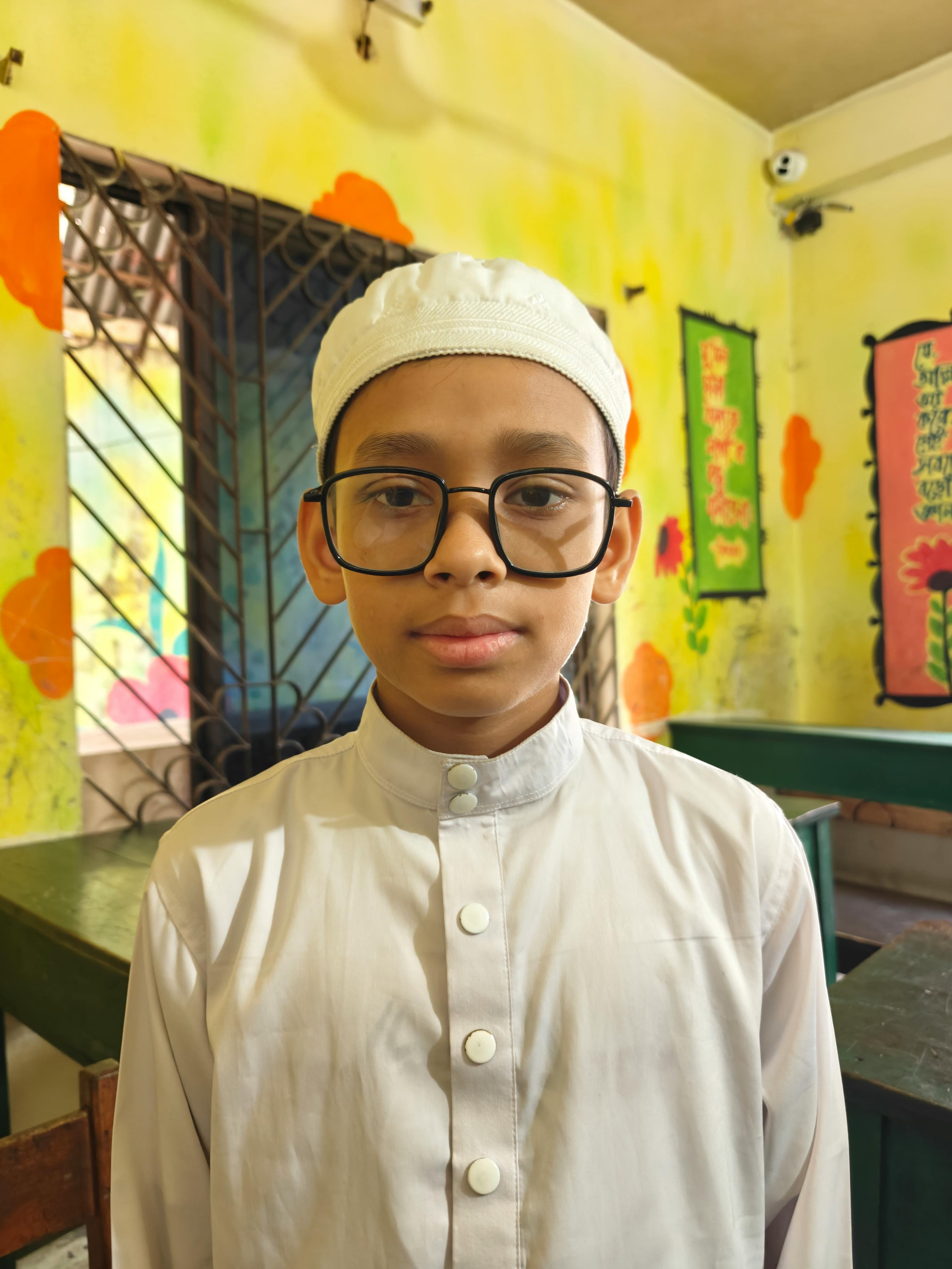 A young boy wearing traditional attire and glasses poses indoors in a colorful room.