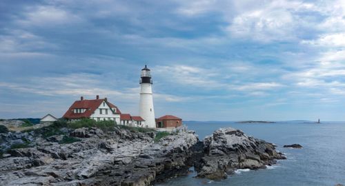 Free Scenic view of a historic lighthouse perched on rocky cliffs with a vast ocean backdrop and cloudy blue sky. Stock Photo