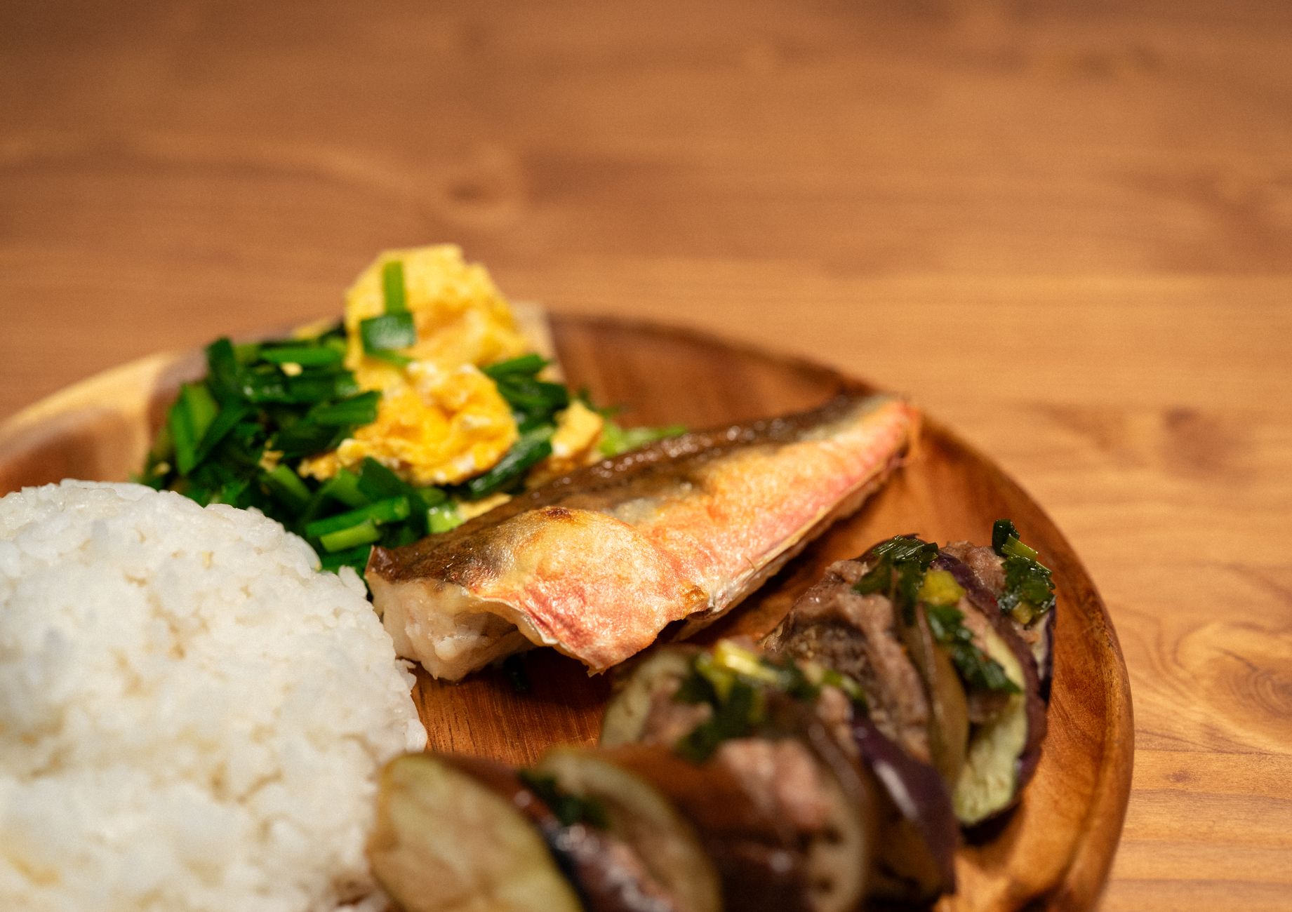 A wooden plate featuring white rice, grilled fish, sautéed vegetables, and scrambled eggs, showcasing Asian cuisine.