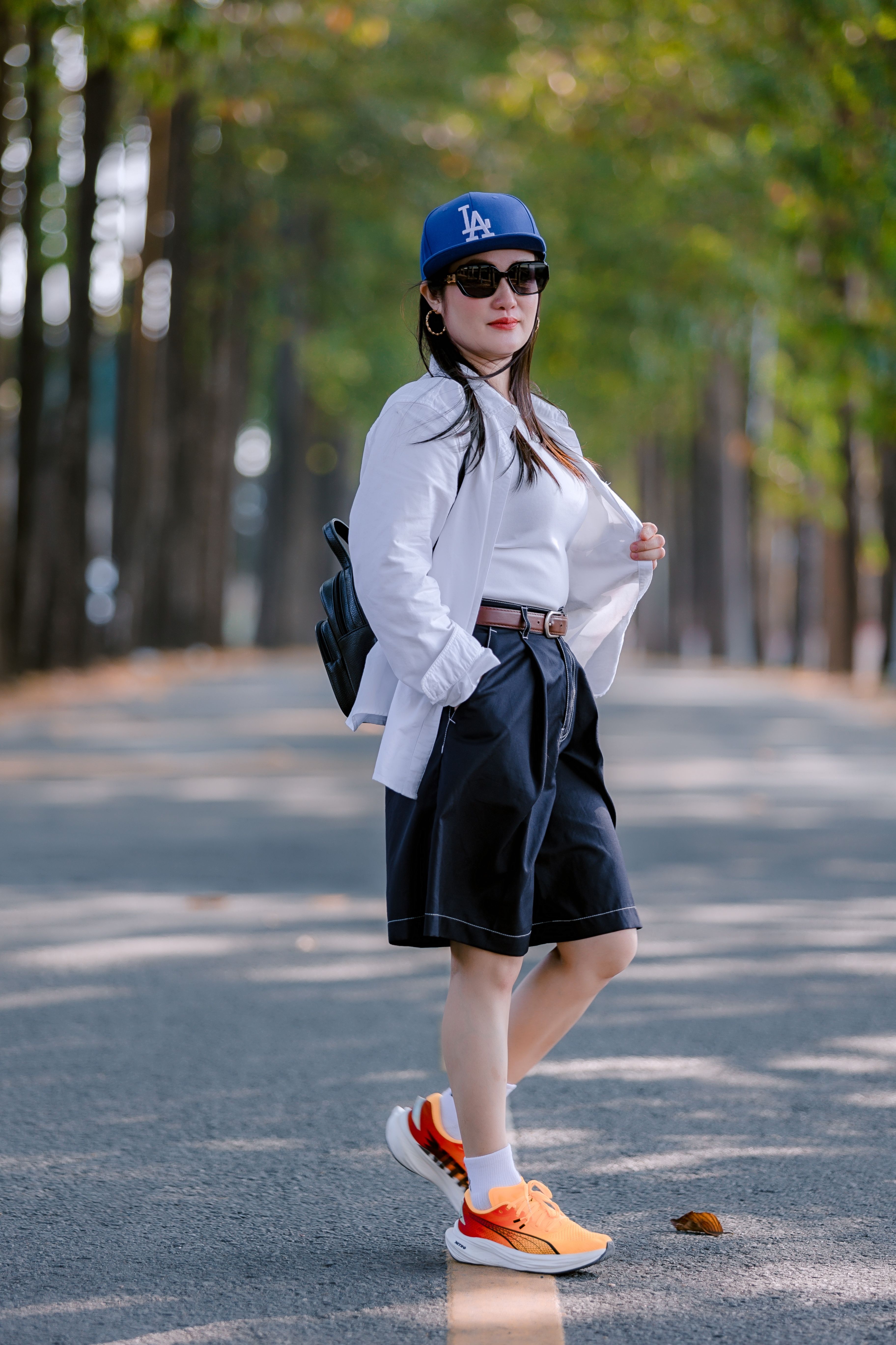 Stylish woman in casual clothing poses on tree-lined street wearing baseball cap and sunglasses.