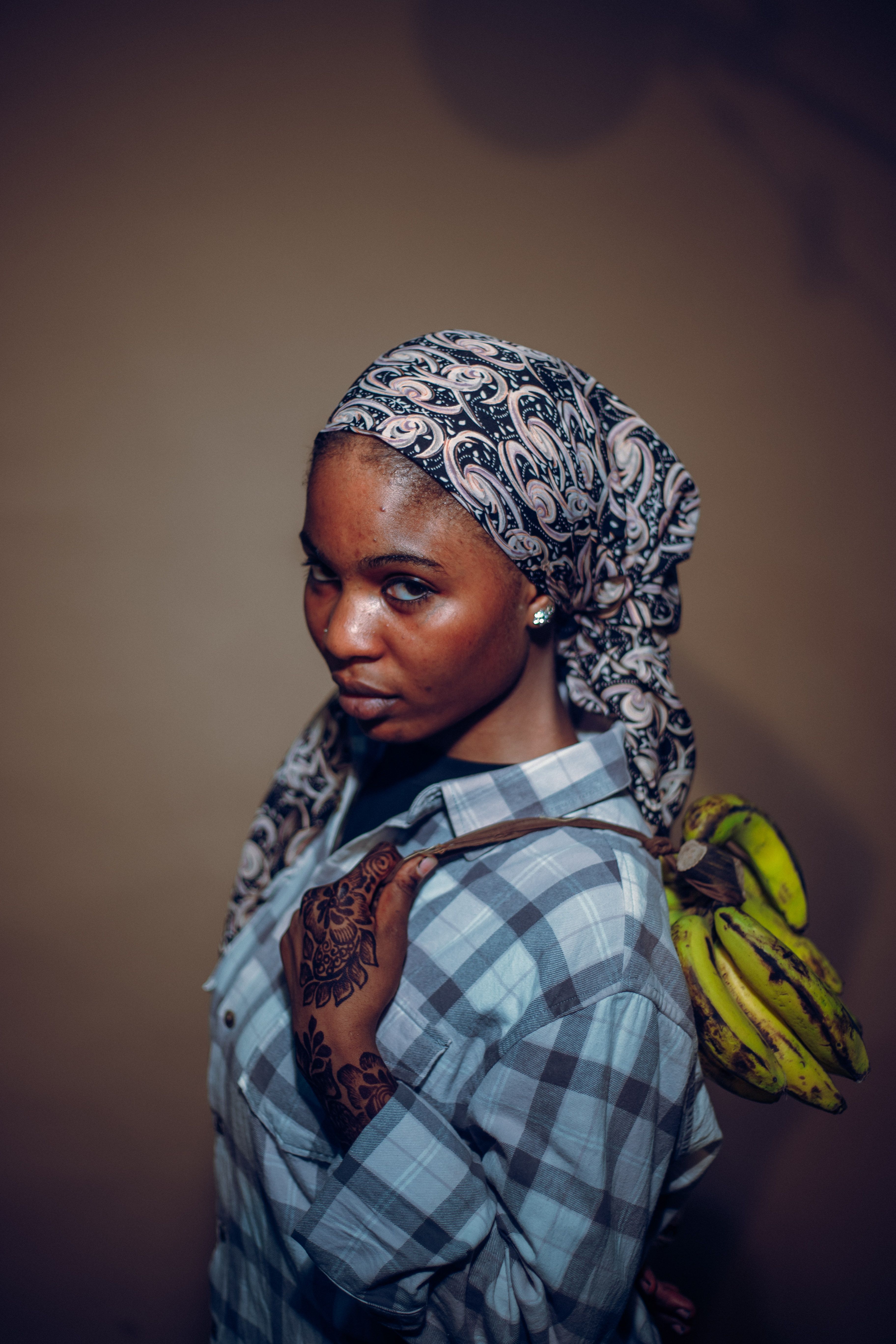 Portrait of an African woman wearing a headscarf and plaid shirt carrying bananas, with henna designs on her hand.