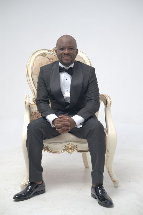 Free Formal portrait of a smiling man in a tuxedo, seated on an ornate chair against a bright background. Stock Photo