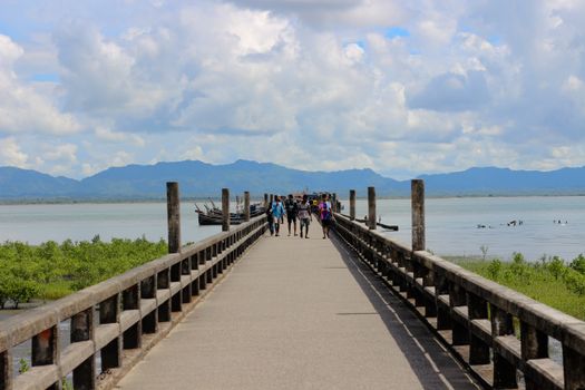 People walking on a wooden pier leading to boats with mountains and blue skies in Bangladesh.