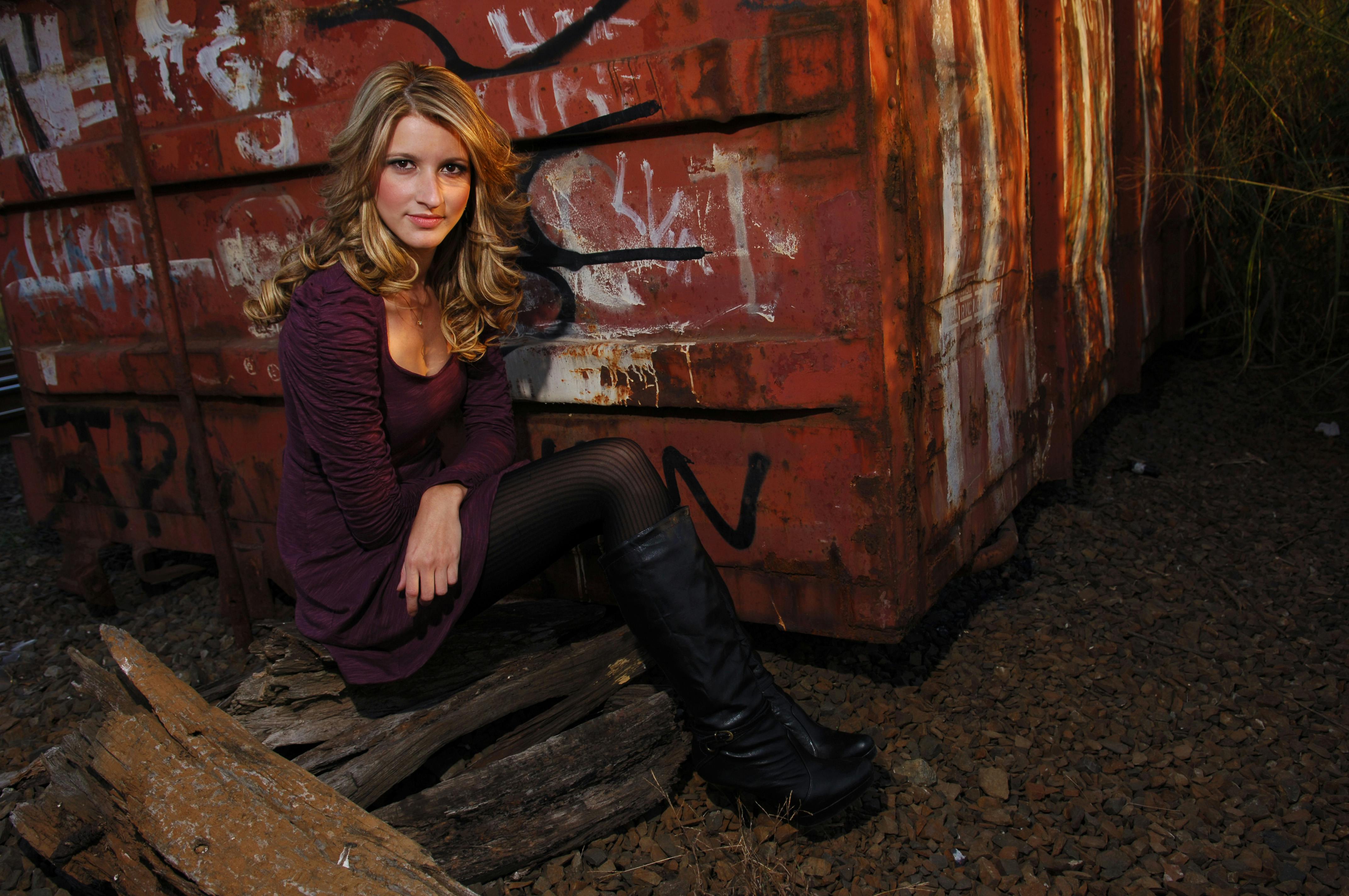 A woman sitting beside a rusty, graffiti-covered container wearing a purple dress and black boots.