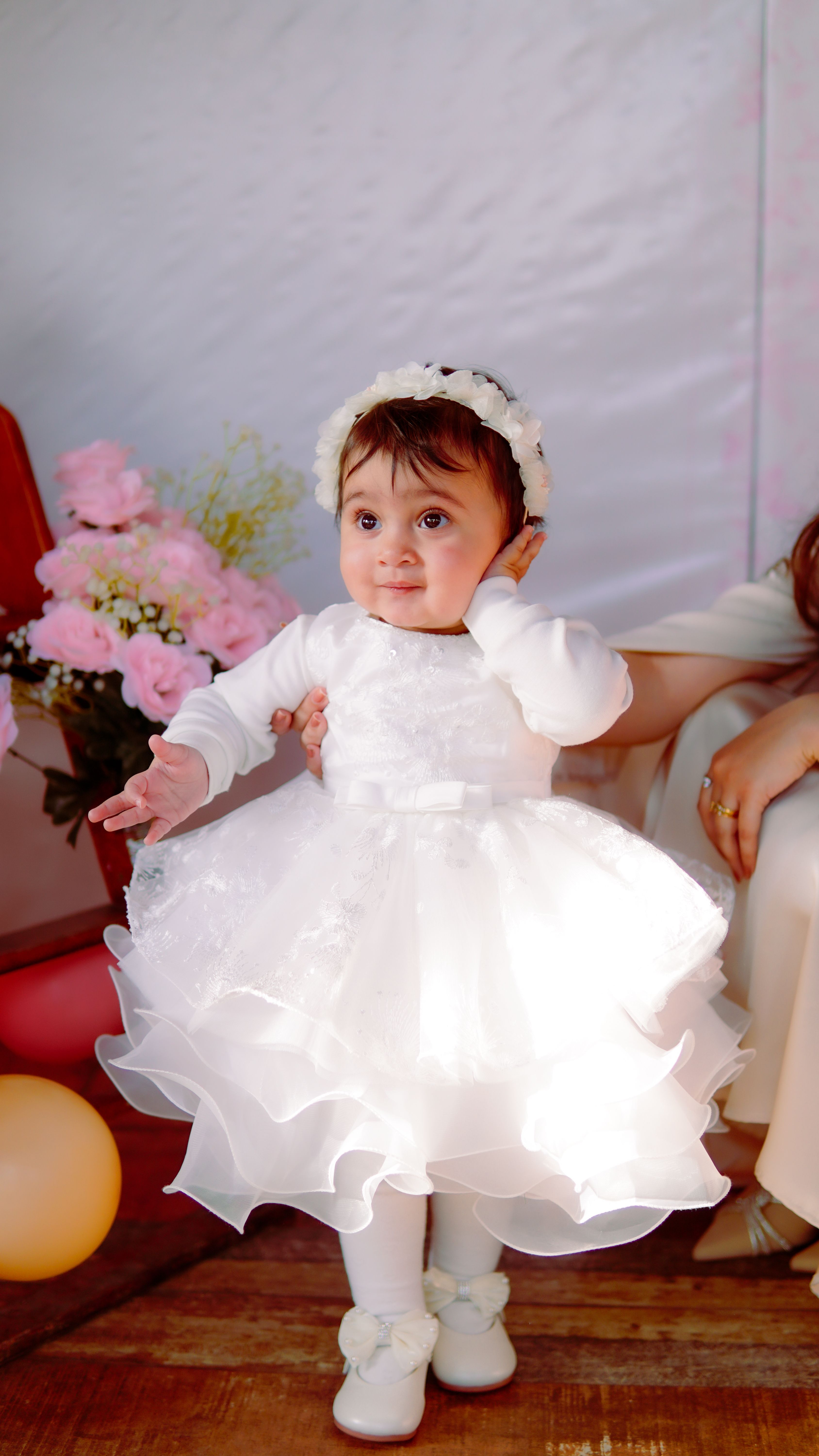 Fotomural Una adorable bebé con un vestido blanco y una diadema de flores en un evento festivo en interiores.