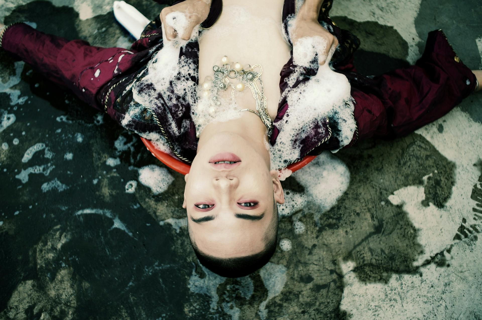 Top view of unrecognizable person touching young male model covered with white foam and lying on concrete ground