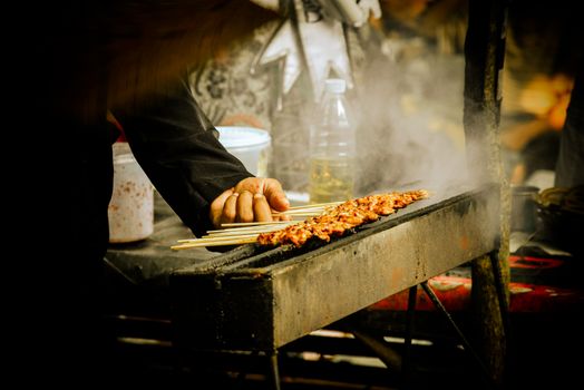 A street vendor grilling skewered meat on an outdoor grill, surrounded by aromatic smoke.