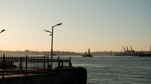 Free A tranquil waterfront scene with a lighthouse and silhouetted industrial cranes during sunset. Stock Photo
