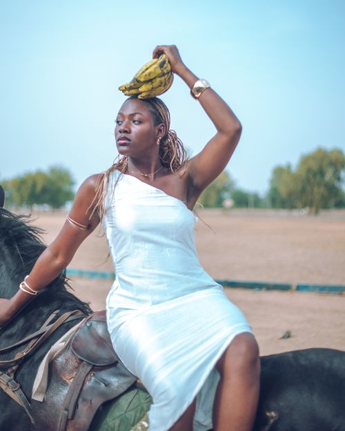 Free Stylish African woman in a white dress posing on a horse with bananas on her head. Stock Photo