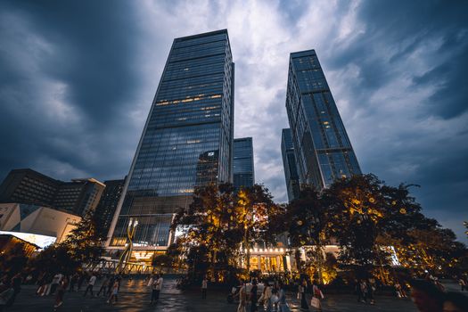 Dramatic view of illuminated skyscrapers against a moody twilight sky, surrounded by bustling urban life.