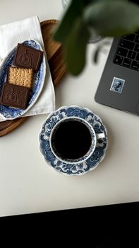 A top-down view of a coffee cup with biscuits and chocolate beside a laptop on a desk.
