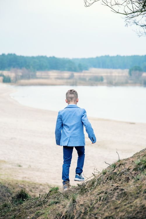 Free Back view of a young boy in a blue suit walking towards a tranquil lakeside in nature. Stock Photo