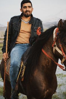 A man in a leather jacket rides a horse through a grassy landscape with distant mountains.