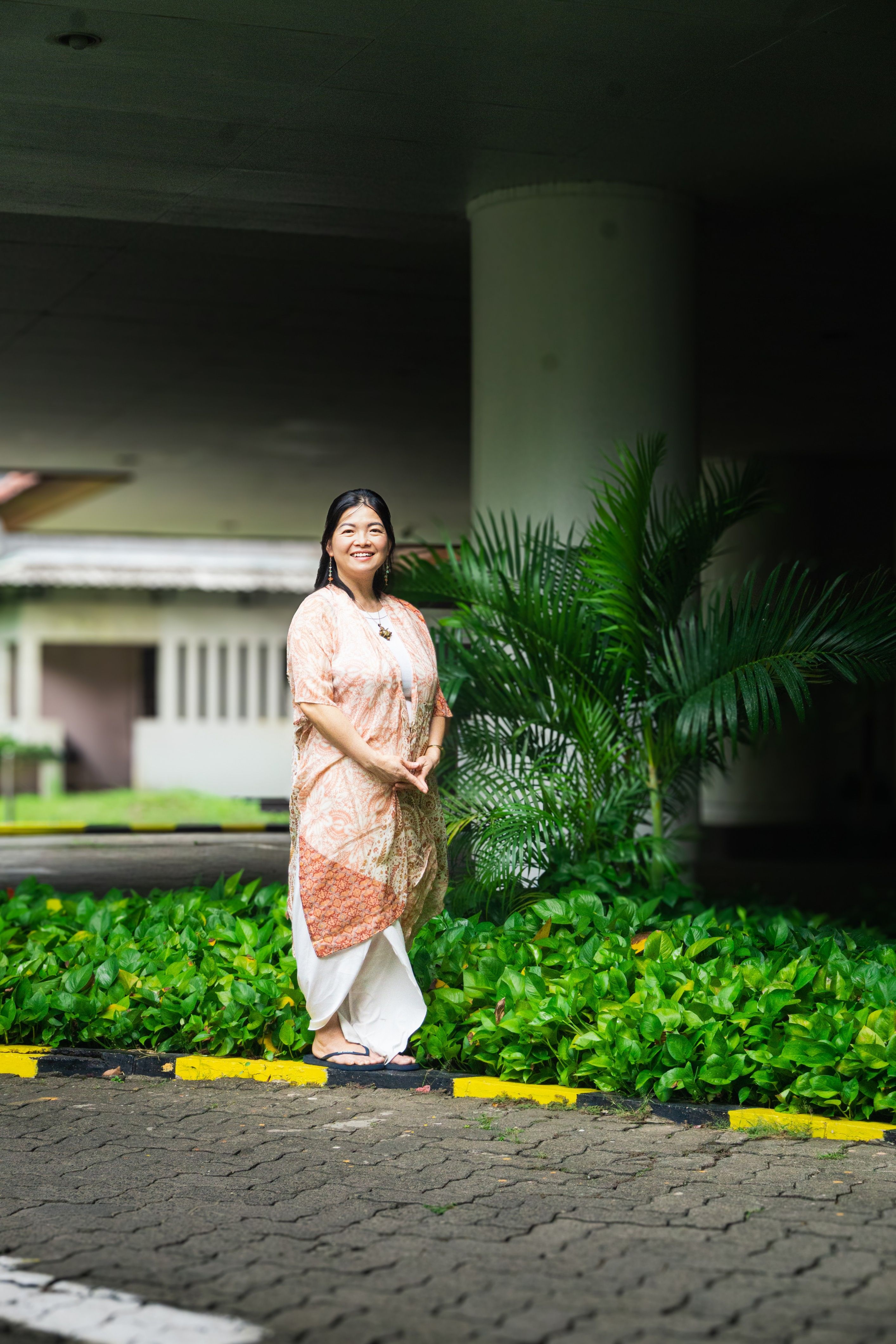 Smiling woman in traditional dress standing outdoors by green foliage.