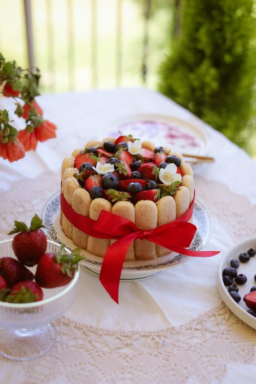 Free Beautiful charlotte cake wrapped with a red ribbon, topped with fresh berries on a lace-covered table outdoors. Stock Photo