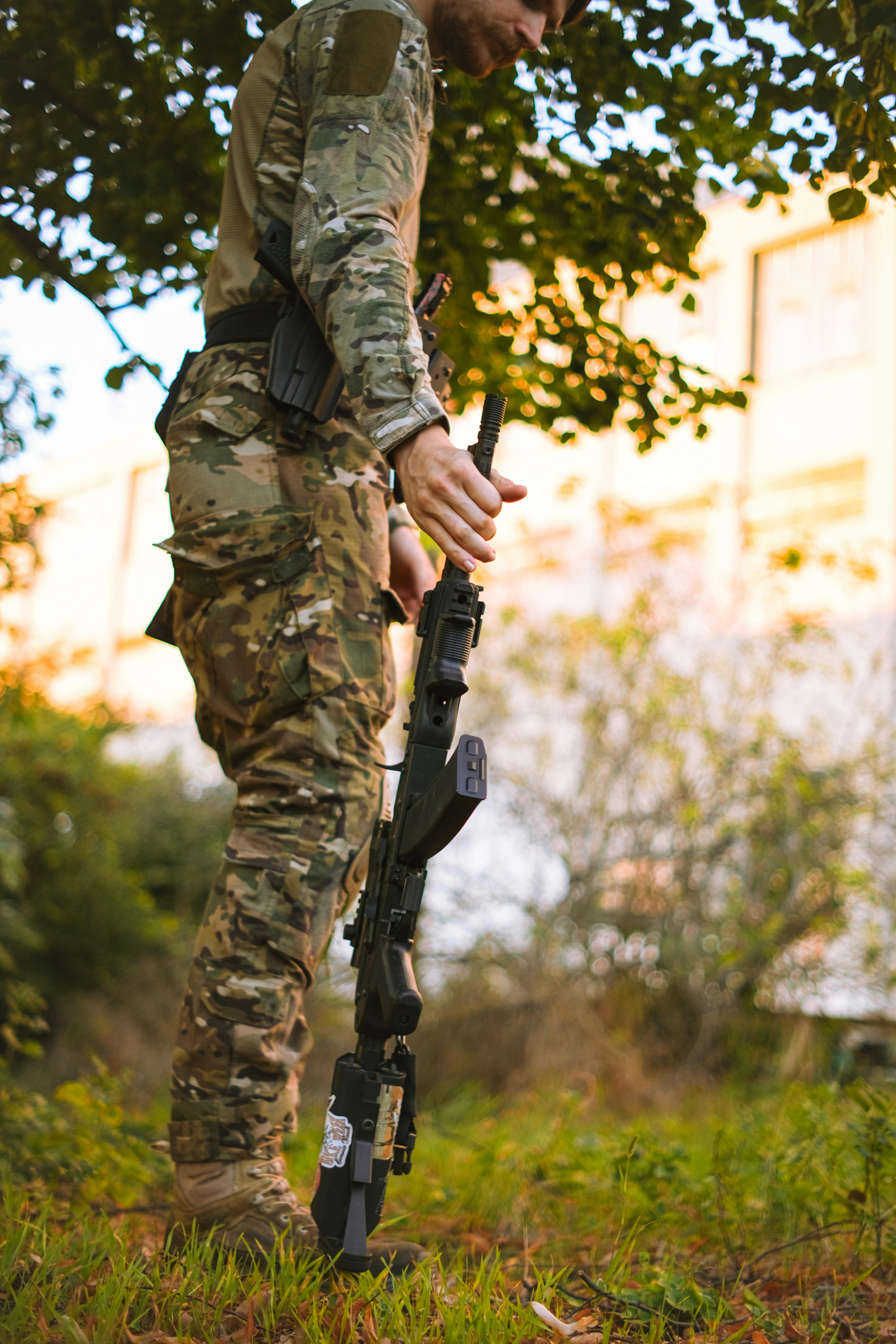 Military personnel in camouflage gear holding a rifle in a natural outdoor setting.