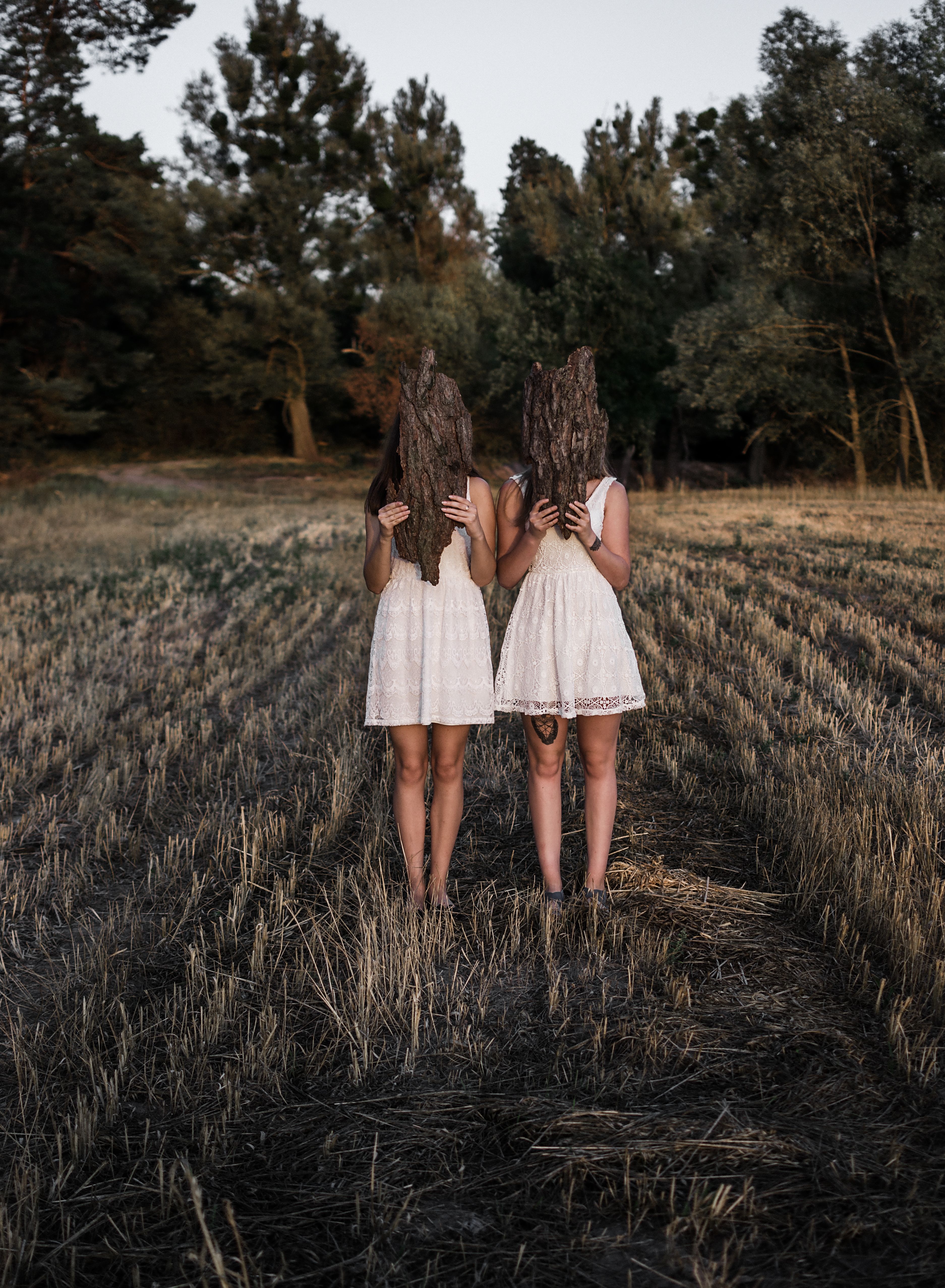 Two women in white dresses hold tree bark in an enigmatic field setting, creating a mystical scene.