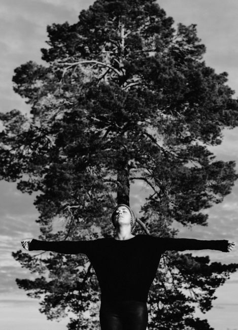 Black and white serene female standing with outstretched arms and closed eyes on background of huge pine tree in woods on sunny day