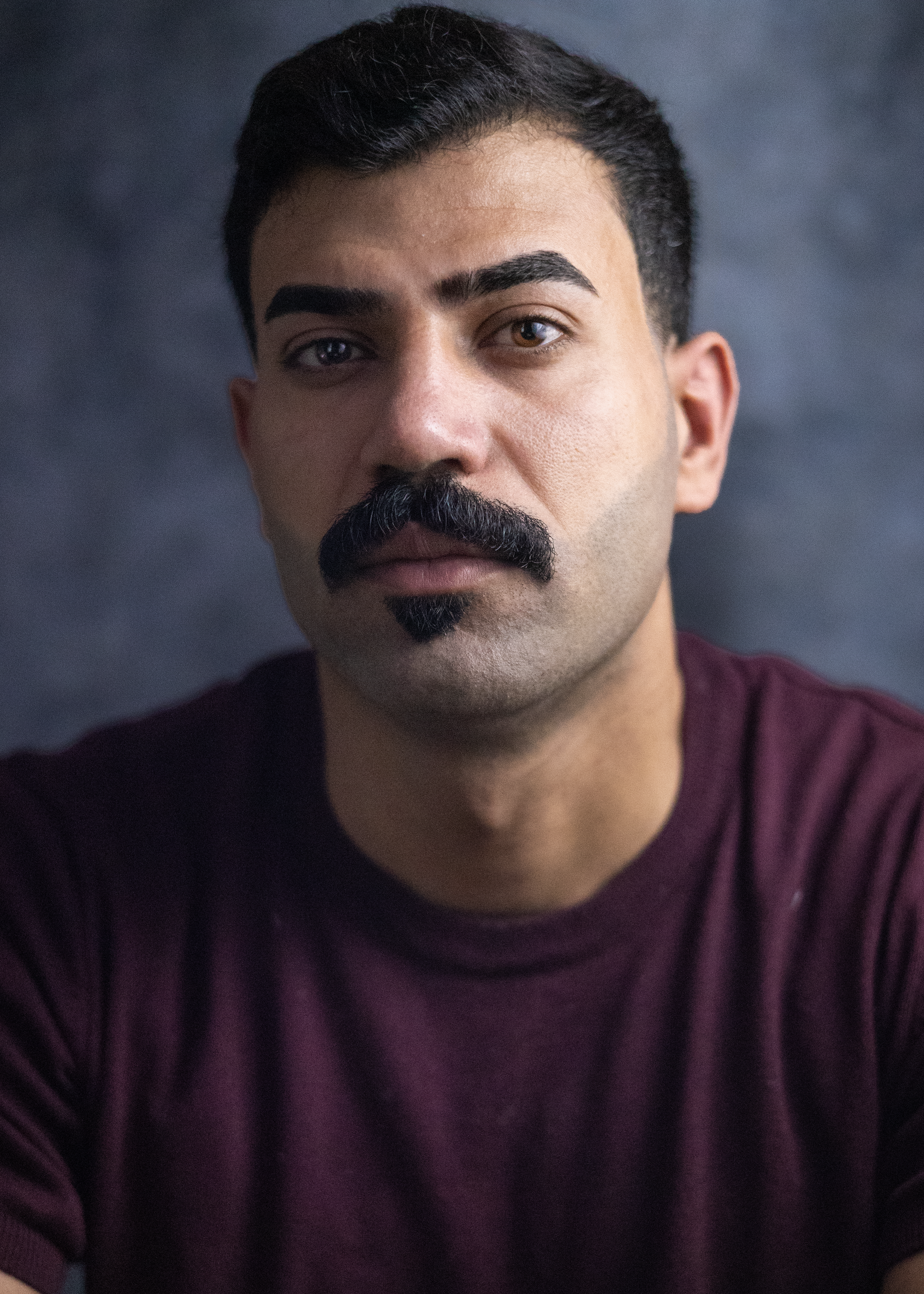 Close-up portrait of a man with a unique mustache, wearing a maroon shirt.