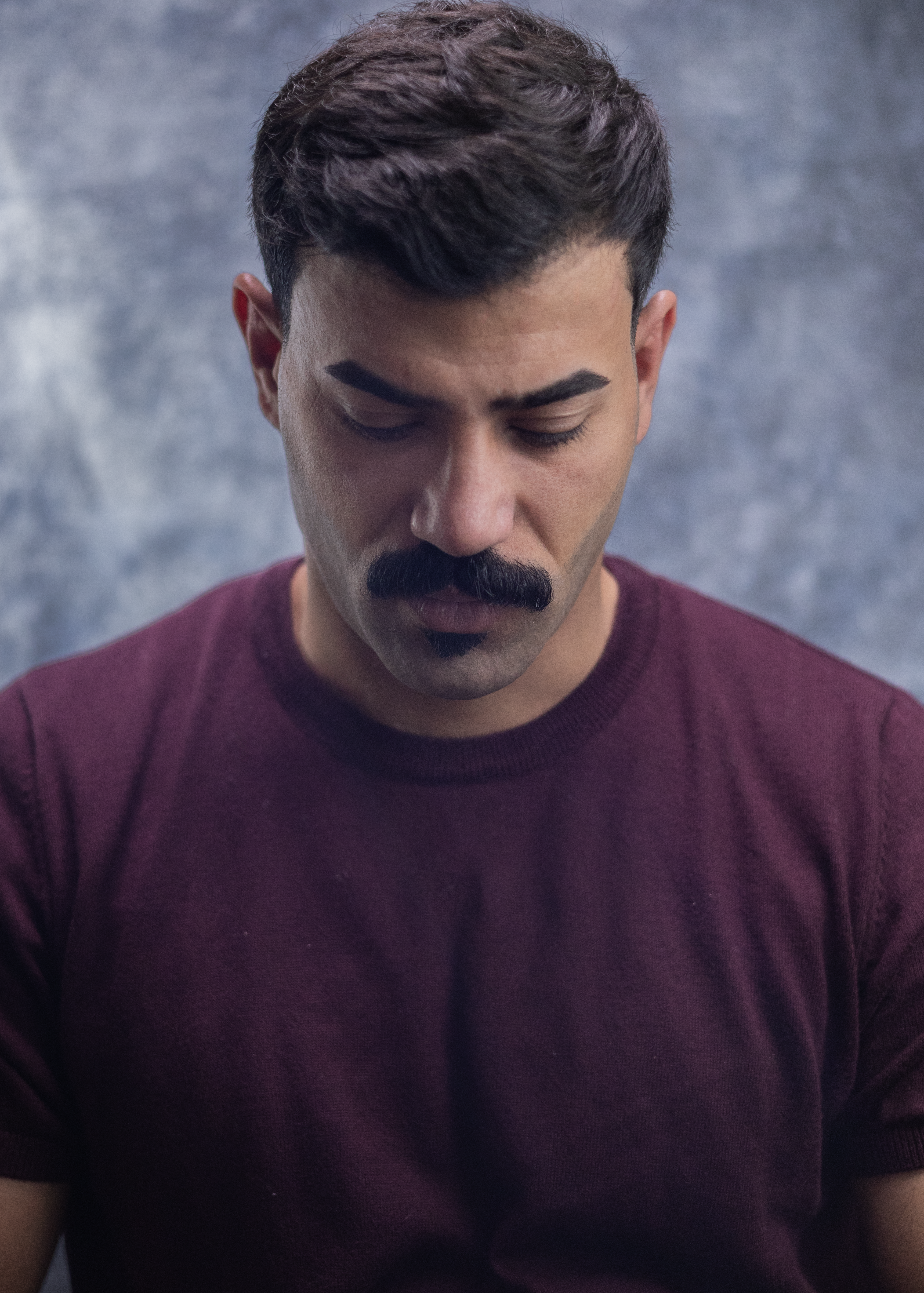 Portrait of a pensive man with a mustache looking down, wearing a maroon shirt.
