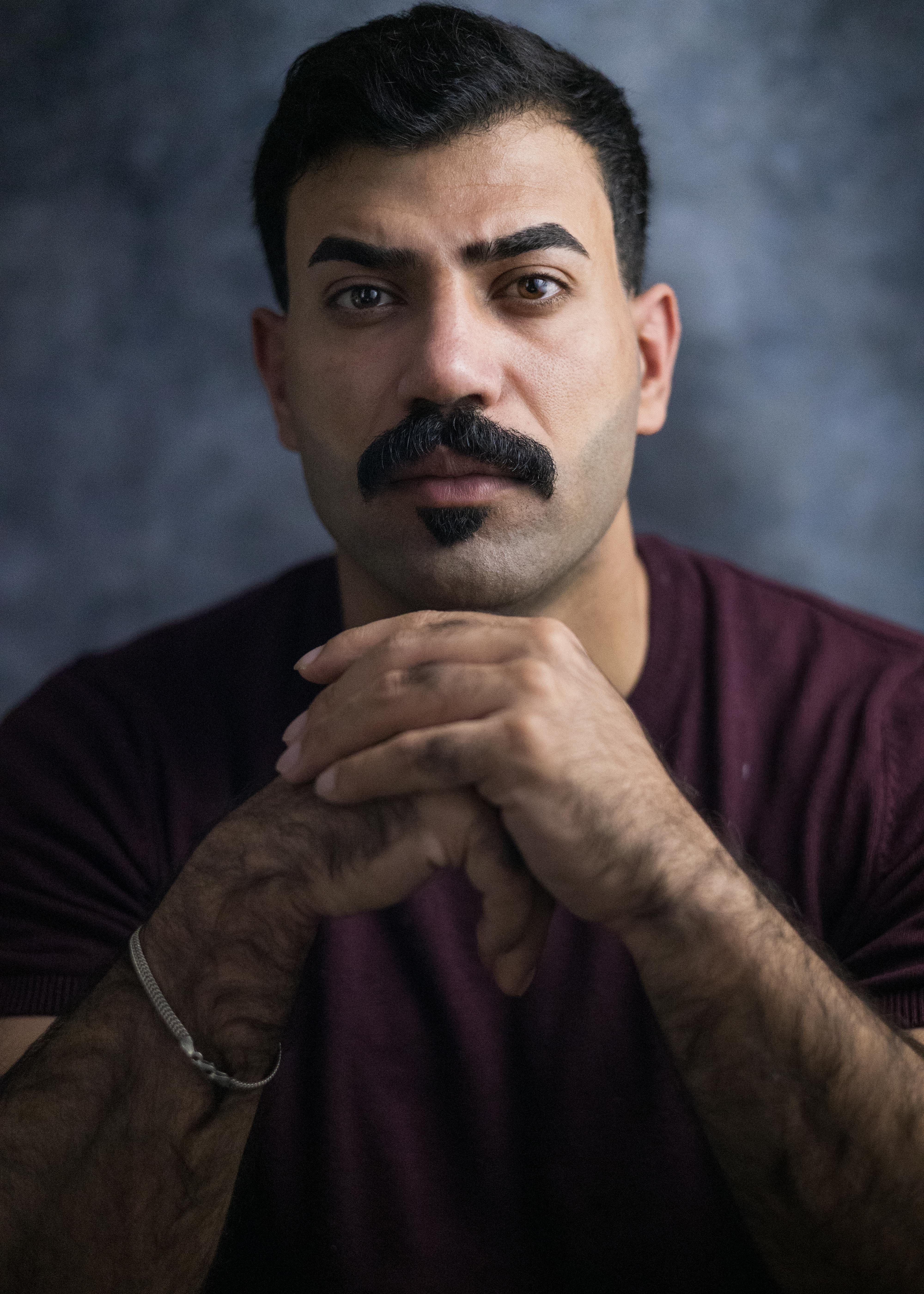 Man with a mustache in a burgundy shirt posing confidently against a neutral backdrop.
