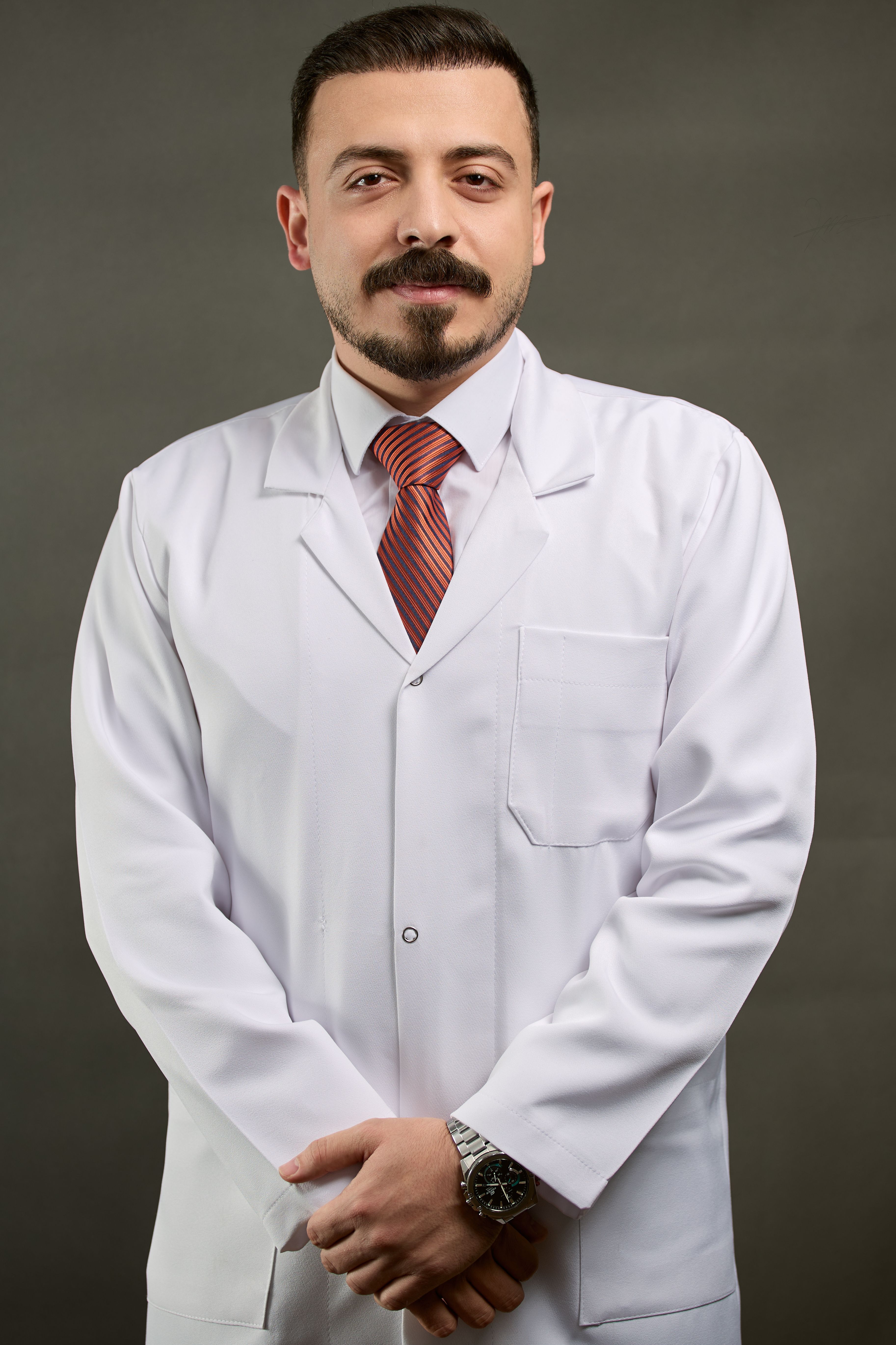 Confident male doctor in a white lab coat with a tie, arms crossed, studio portrait.