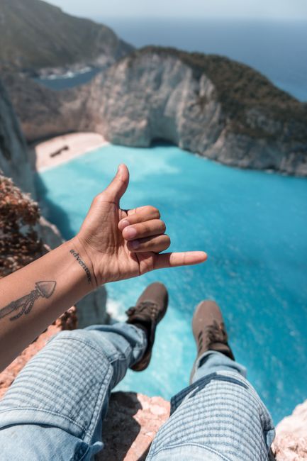 Person with tattoo gives hand gesture overlooking Zakinthos cliffs and sea.
