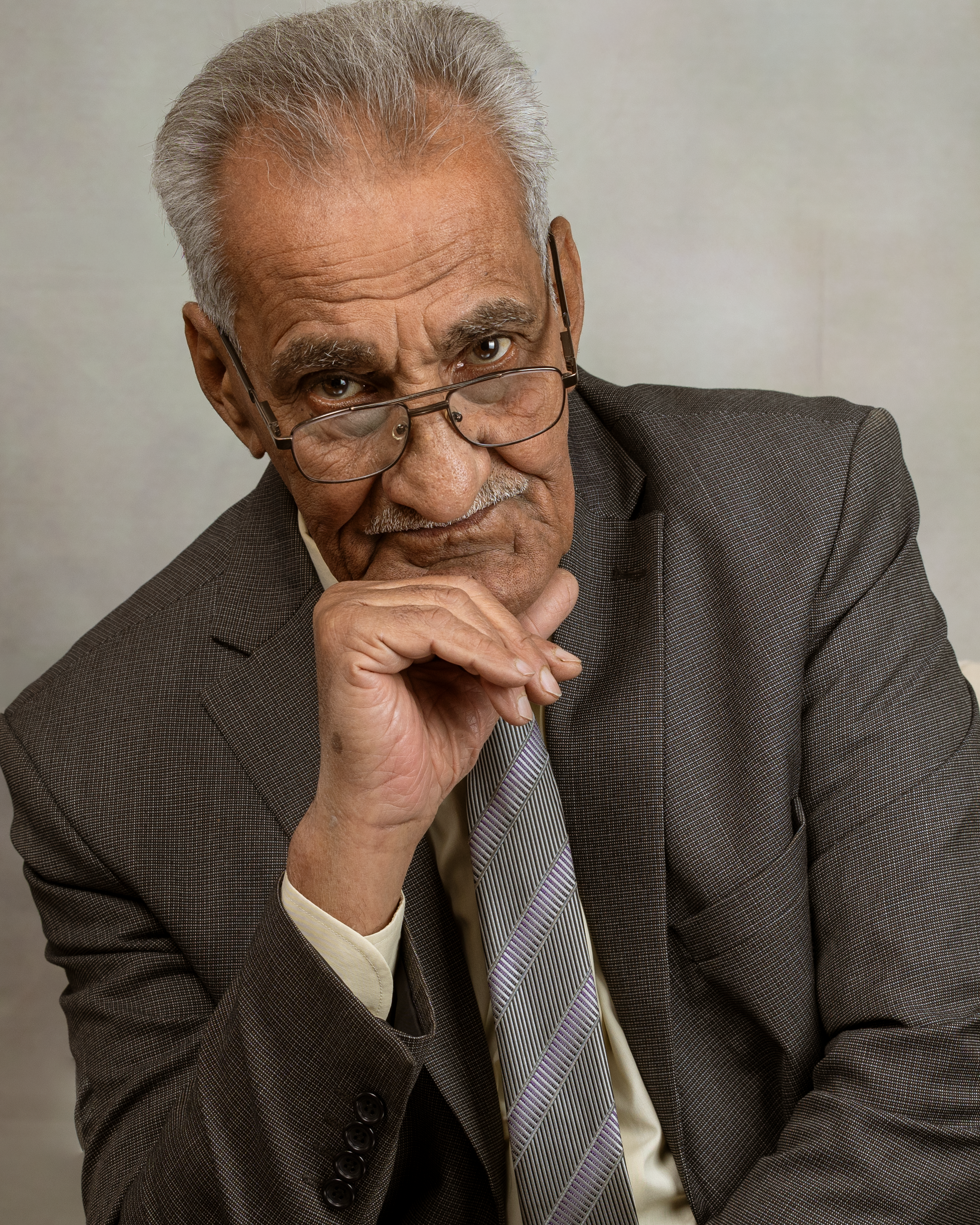 Elderly man in a suit with glasses, posing thoughtfully against a neutral background.