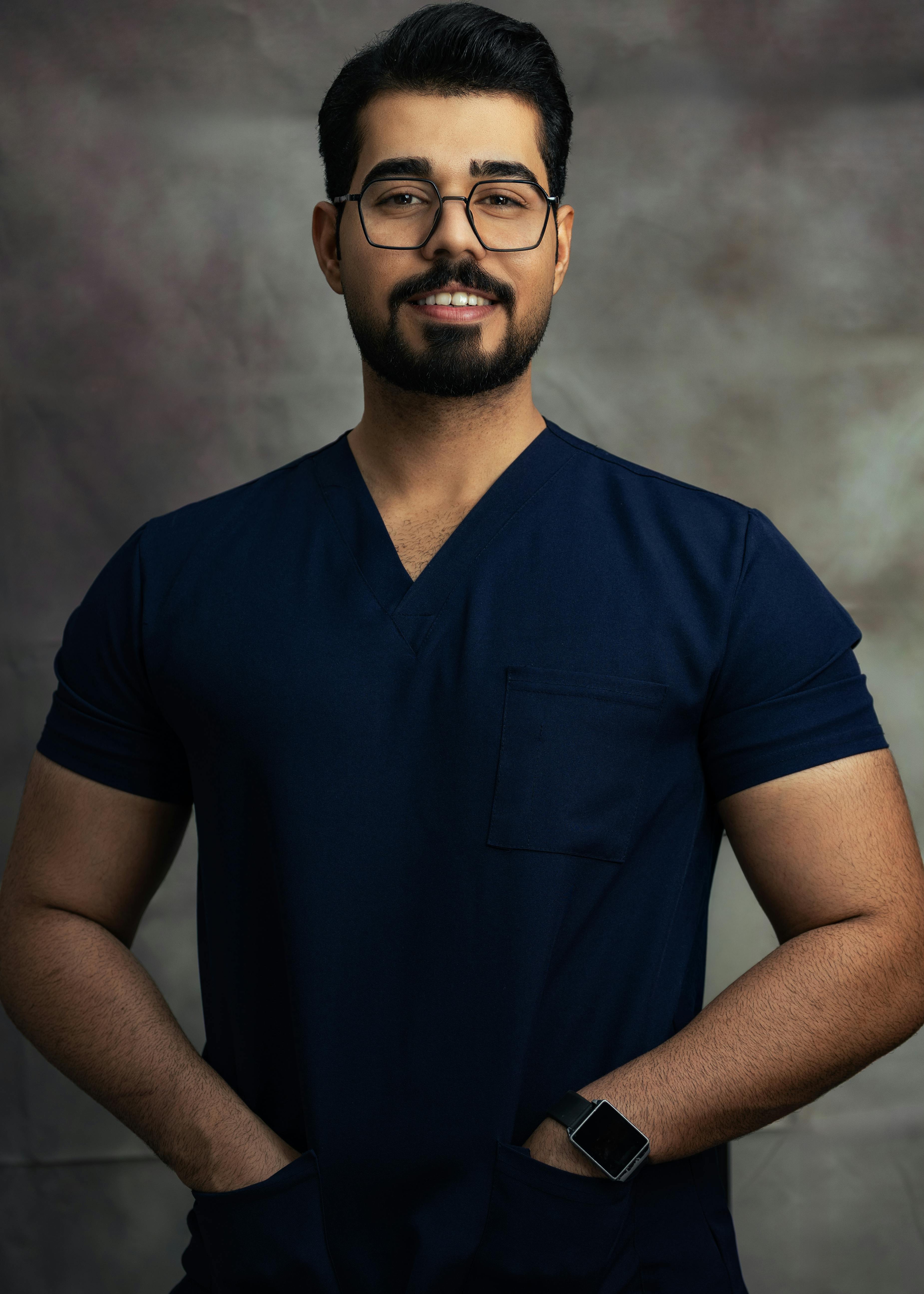 Smiling man wearing blue scrubs, standing confidently indoors with hands in pockets.