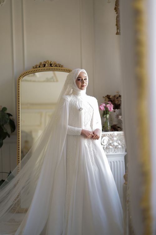 Free Bride in elegant white wedding gown standing indoors with a mirror backdrop, exuding grace and beauty. Stock Photo